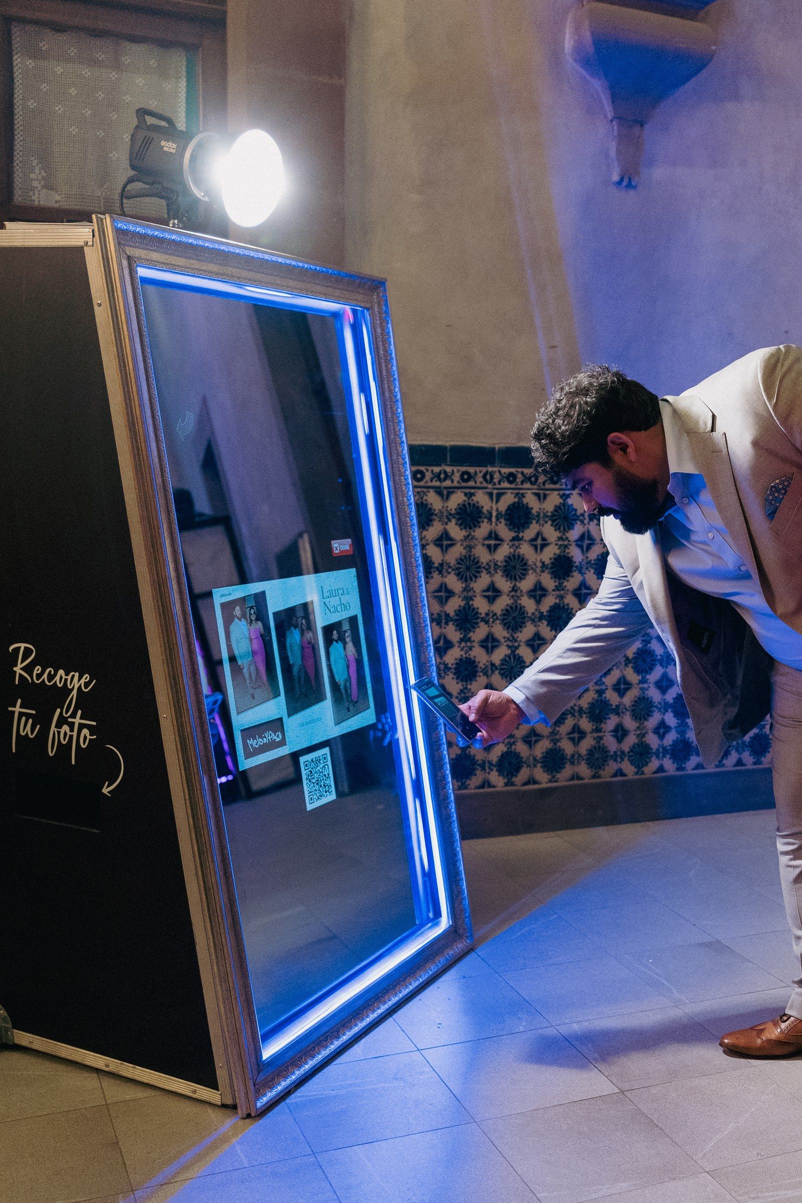 Invitado escaneando el QR del espejo mágico Pikibooth para descargar su foto, con luz de estudio y letrero Recoge tu foto
